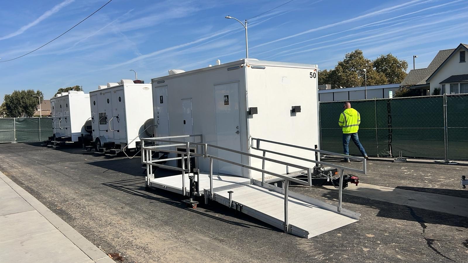 A Lavatory accessible restroom trailer with a long aluminum ramp on a paved job site lot, with more trailers lined up behind it and a worker in a safety vest walking past