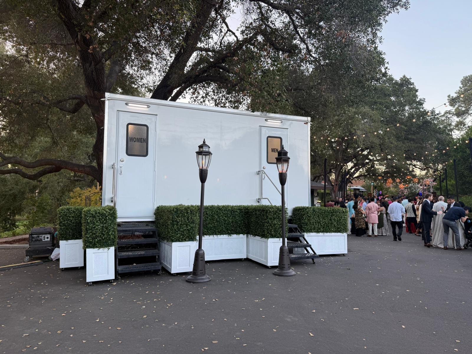 A Lavatory restroom trailer with women's and men's doors, dressed with boxwood planters and lamp posts at an evening reception under large oak trees, with guests gathered nearby