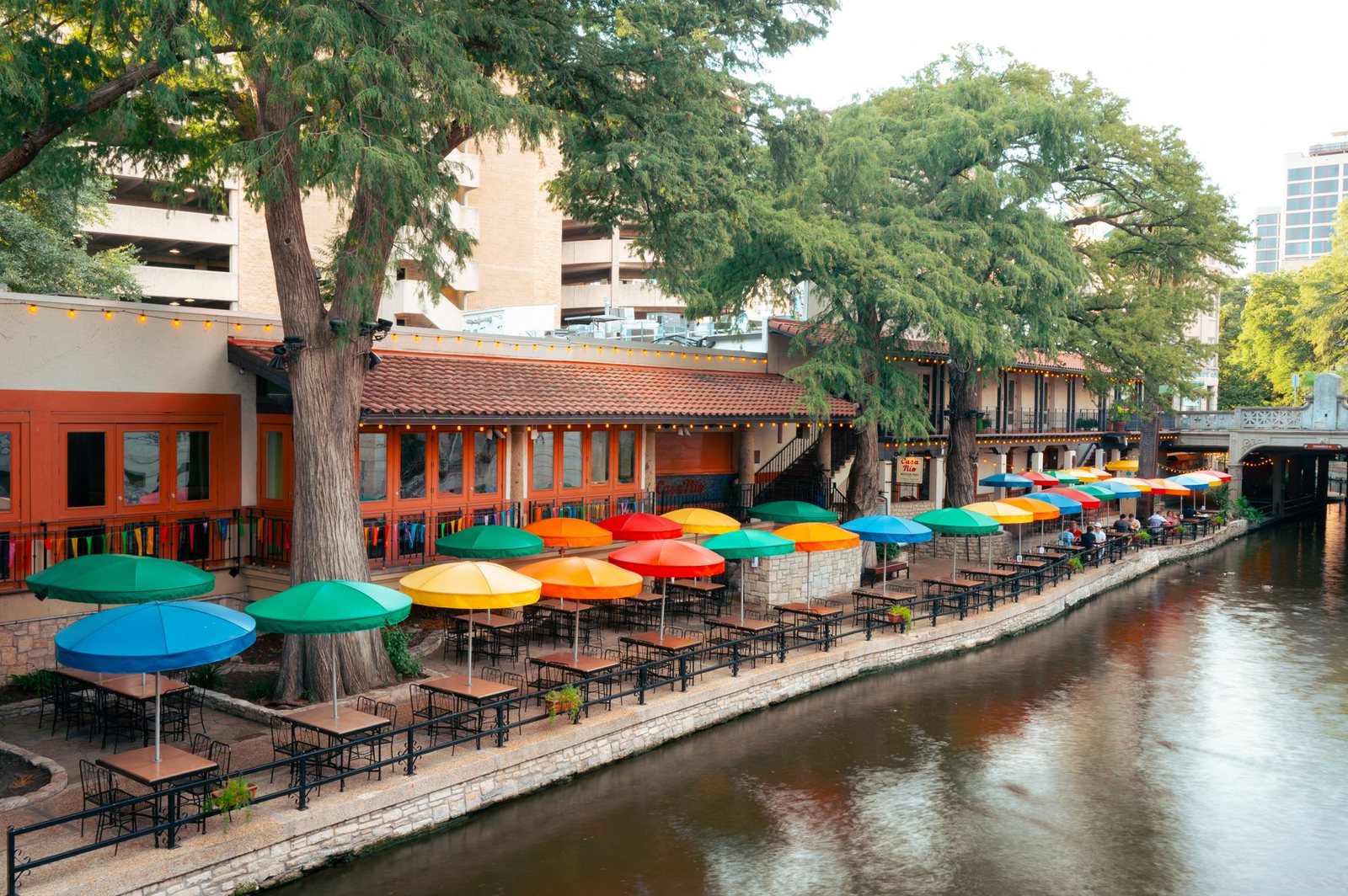 Rows of bright patio umbrellas over riverside restaurant tables along the San Antonio River Walk, shaded by cypress trees